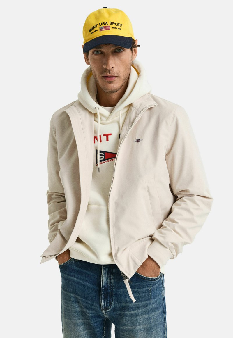 Chaqueta beige con cremallera, sudadera con capucha con logo, jeans azules y una gorra amarilla y azul marino con texto. Textura suave, diseño casual.