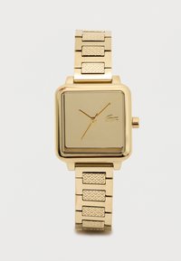 Kultakylkinen neliömäinen kello, jossa on tekstuurilla varustetut linkit, minimalistinen kellotaulu, jossa on kolme kultakättä ja pieni krokotiililogo kello kolmessa.