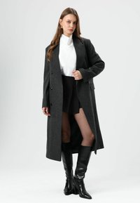 Grijze wollen jas met een lange, rechte snit, voorzien van zes knopen en zijzakken. Gecombineerd met een witte coltrui en een zwarte minirok.