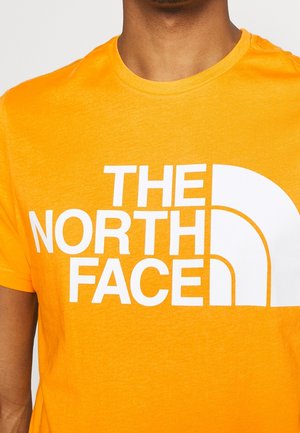 Jasnopomarańczowa bawełniana koszulka z okrągłym dekoltem. Posiada duży biały napis "THE NORTH FACE" oraz zakrzywione logo. Gładka tekstura.