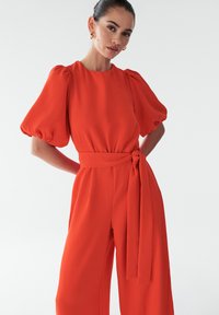 Punane kombinesoon puffsevartega, vöökoha ja laia säärekujundusega. Sujuv kangas puhta ja modernse silueti jaoks.