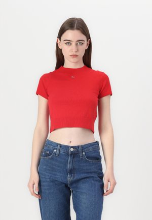 Top a maglia corto rosso con maniche corte e collo alto. Dettaglio distintivo di un piccolo logo sul petto. Abbinato a jeans in denim a vita alta.
