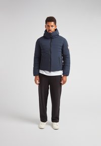 Marineblå pufferjakke med høj krave, lynlås foran og to sidelommer. Iført over en hvid skjorte, parret med sorte bukser og hvide sneakers.