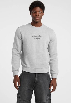 Sivý sveter s okrúhlym výstrihom, so žebrovanými manžetami a spodným lemom. Na hrudi sa nachádza čierny logotyp a emblém "Guess Jeans". Kombinovaný s tmavými cargo nohavicami.