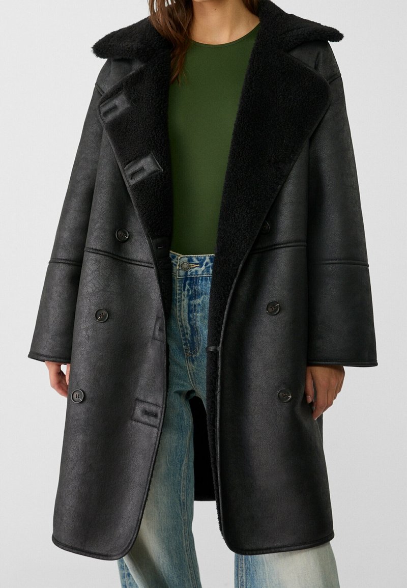 Personne portant un long manteau noir en peau lainée avec de larges revers sur un haut vert et un jean bleu, visible du cou aux genoux.