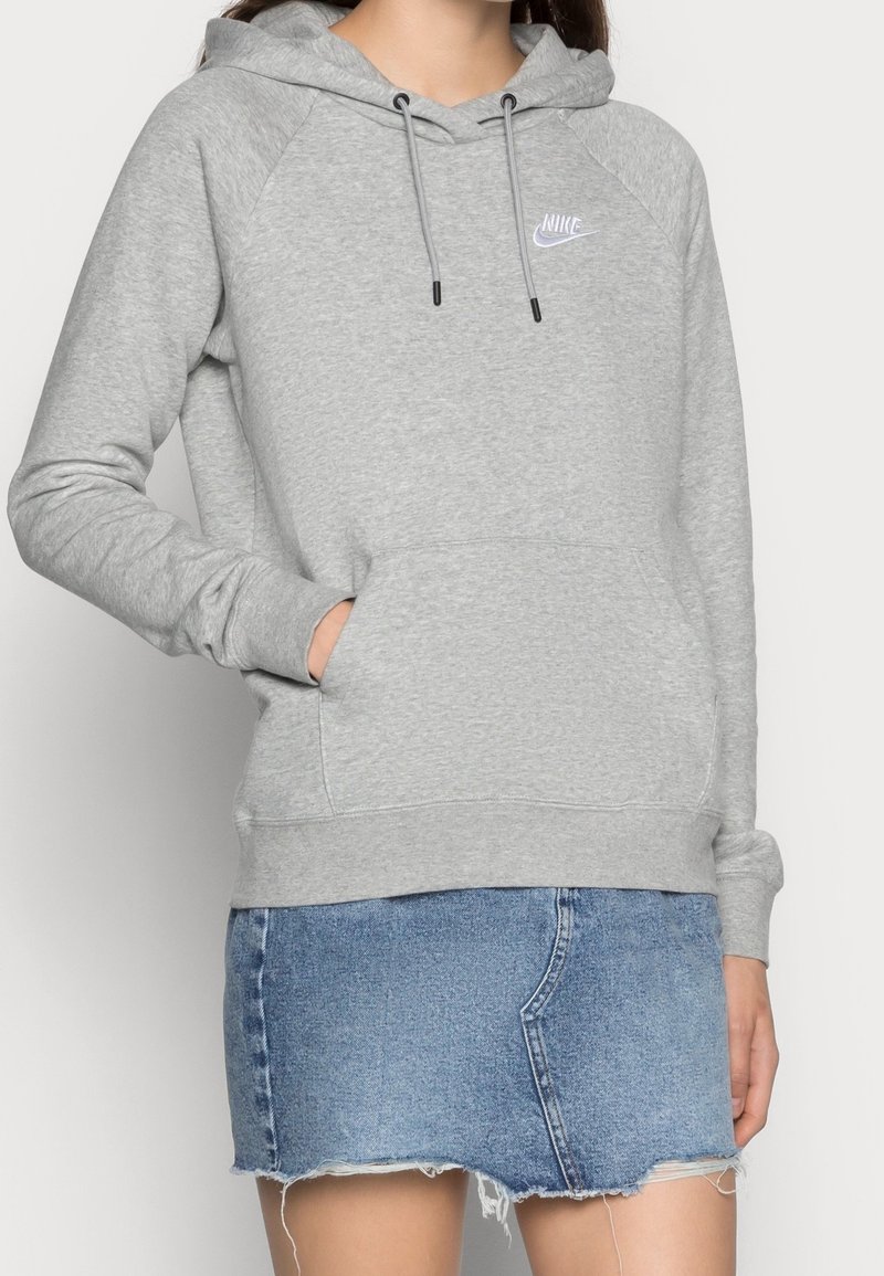 Femme portant un sweat à capuche Nike gris clair avec poche avant et jupe en denim bleu effilochée, main dans la poche, vue du cou jusqu'à mi-cuisse.