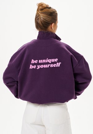 Femme aux cheveux blonds attachés en chignon porte une veste violette avec le texte rose « be unique be yourself » dans le dos et un pantalon blanc sur un fond uni.