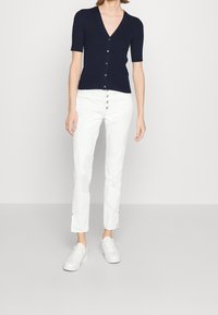 Marinblå ribbad kortärmad cardigan med V-ringning och knäppning fram, matchad med vita slim-fit byxor med dekorativa knappar vid midjan.