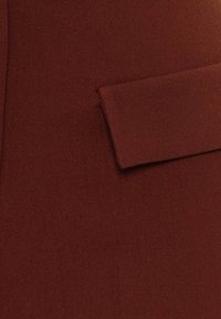 Roestkleurige stof met een gladde textuur en een enkele flapzakdetail. Het materiaal lijkt lichtgewicht en op maat gemaakt.