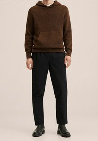 Sweat à capuche marron duveteux avec une poche avant, associé à un pantalon plissé noir. Le mannequin porte des bottines noires. Fond neutre.