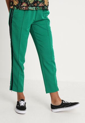 Persoon die groene cropped broek draagt met zwarte zijstrepen en zwarte veterschoenen op een effen lichte achtergrond.