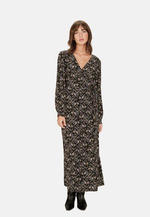 Robe longue noire avec un motif floral, décolleté en V, manches longues et taille à nouer. Tissu doux et design fluide. Portée avec des bottes noires à la cheville.