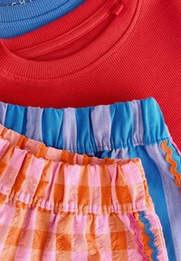 Tres prendas apiladas: un suéter rojo con estrías, una blusa de rayas azul y una falda con volantes multicolor con un patrón de cuadros rosa-naranja y acentos.
