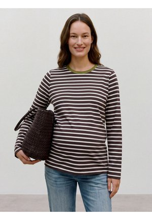 Femme souriante portant un t-shirt à manches longues rayé marron et blanc, un jean bleu, et tenant un grand sac à main tressé sombre sur son bras gauche.