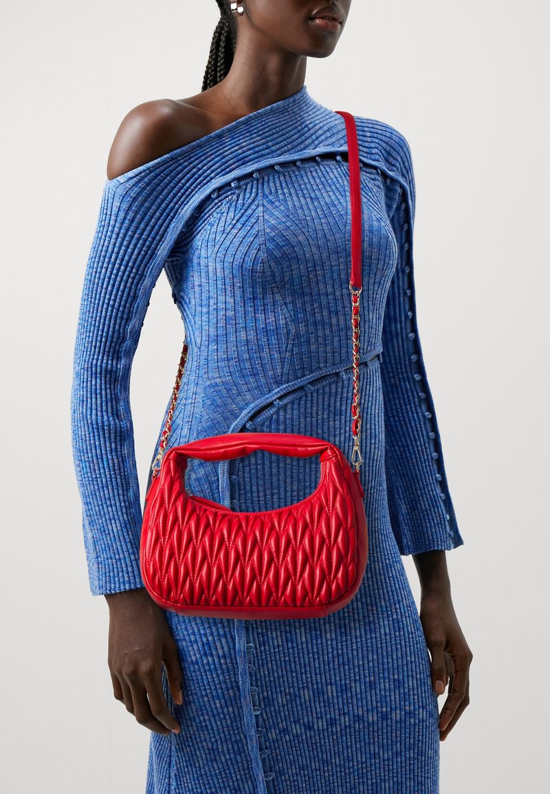 Femme portant une robe bleue côtelée à épaules dénudées avec des détails de boutons, portant un sac à main rouge matelassé avec une bandoulière chaîne sur l'épaule.