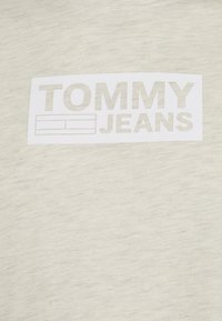 Světle béžové bavlněné tričko s centrálním bílým obdélníkovým logem, na němž je nahoře nápis "TOMMY" a dole "JEANS" v šedém písmu. Měkká textura.