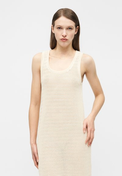 Γυναίκα με ίσια καστανά μαλλιά φοράει αμάνικο κρεμ φόρεμα πλεχτό με βελονάκι, στέκεται μπροστά σε απλό φωτεινό φόντο.