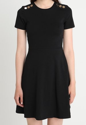 Femme portant une robe noire à manches courtes avec un col rond et des boutons dorés décoratifs sur les deux épaules, sur un fond uni.