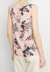 Top senza maniche rosa con stampa floreale verde scuro e rossa e motivi a farfalla, chiusura a bottone a chiave sul retro, indossato con pantaloni beige chiaro.