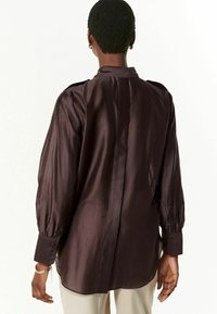 Chemise en soie violet foncé avec un col montant, des manches froncées et un ourlet incurvé. Elle comporte des boutons aux poignets et présente une texture lisse et brillante.