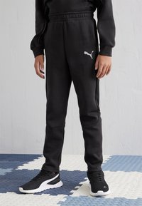 Pantalones de chándal negros hechos de una tela suave con cintura elástica, con un logo de Puma blanco en el muslo izquierdo. Combinados con zapatillas deportivas negras.