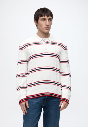 Junger Mann, der einen weißen Strickpullover mit roten, schwarzen und blauen horizontalen Streifen und blaue Jeans trägt, steht vor einem einfarbigen Hintergrund.