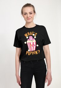 Czarna koszulka z krótkim rękawem z barwną grafiką przedstawiającą niedźwiedzia w pudełku z popcornem w czerwone paski, z napisem "CO SŁYCHAĆ?" nad obrazkiem.