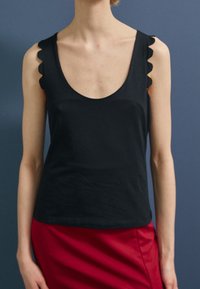 Černý bezrukávový top s hlubokým kulatým výstřihem, s vlnitými okraji na ramínkách. Materiál je hladký a přiléhavý.