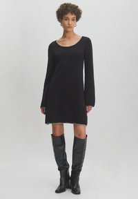 Schwarzes Langarmkleid mit Rundhalsausschnitt, verziert mit dekorativen Nähten am Saum und an den Bündchen, kombiniert mit hohen schwarzen Lederstiefeln.