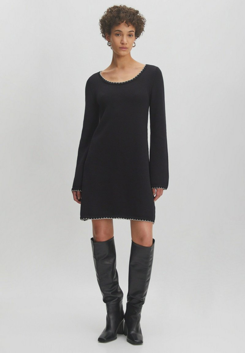 Schwarzes Langarmkleid mit Rundhalsausschnitt, verziert mit dekorativen Nähten am Saum und an den Bündchen, kombiniert mit hohen schwarzen Lederstiefeln.