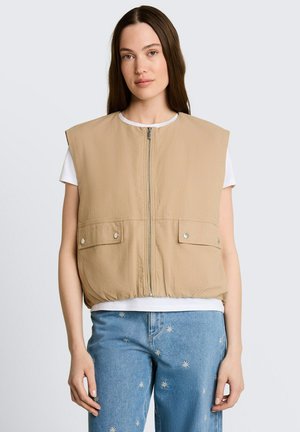 Frau mit langen braunen Haaren, die eine beige ärmellose Reißverschlussweste über einem weißen T-Shirt und blaue Jeans mit kleiner weißer Blumenstickerei trägt.