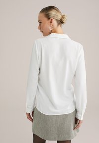 Una donna con capelli biondi raccolti in un chignon indossa orecchini a cerchio d'argento, una maglietta bianca a maniche lunghe e una gonna grigia a spina di pesce, di spalle.