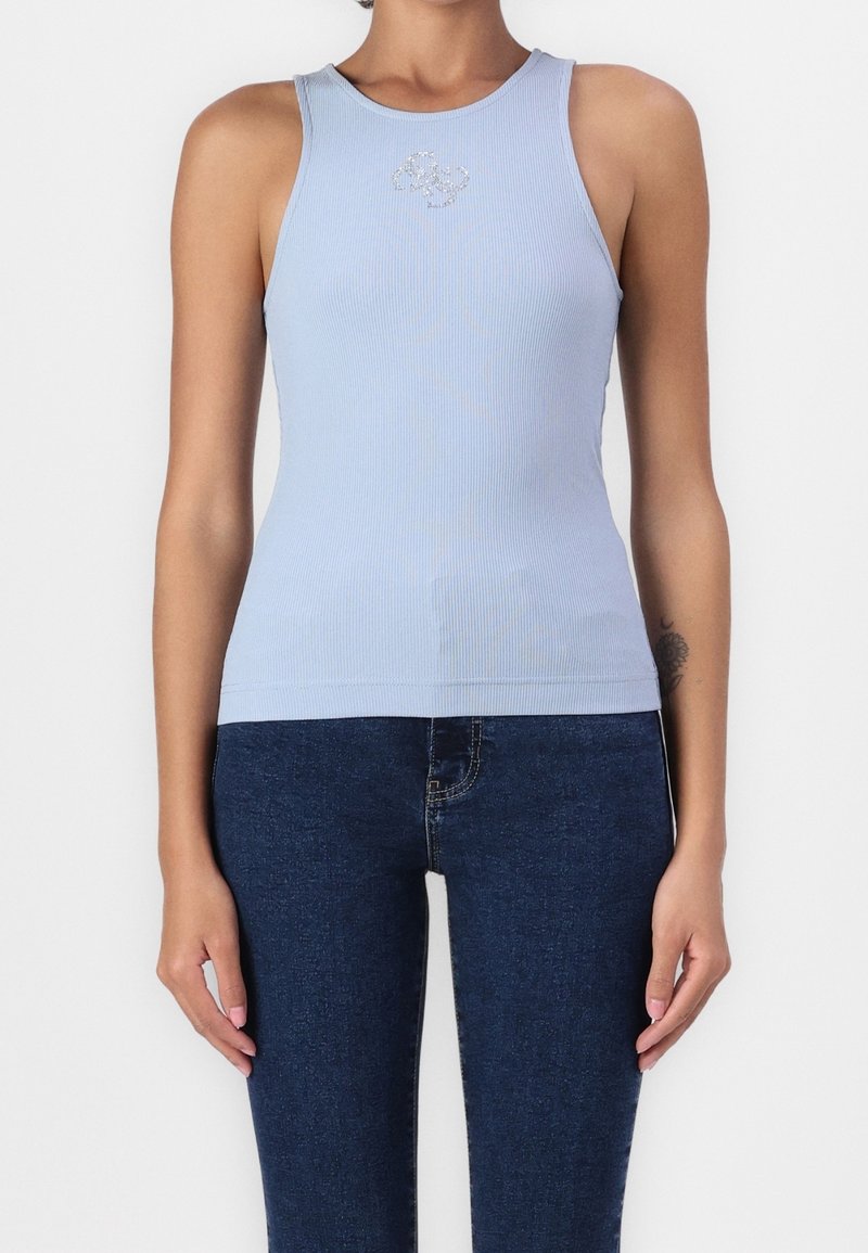 Femme portant un haut bleu clair sans manches à côtes avec un petit motif en strass près de l'encolure et un jean bleu foncé.