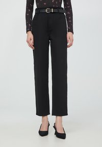 Pantaloni neri a vita alta con taglio dritto, dotati di una cintura con fibbia dorata. Tessuto liscio e tasche frontali. Abbinati a tacchi neri.