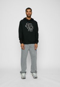 Sort hoodie med hvid grafik på forsiden, grå jeans og lysegrå sneakers. Hoodien har snore og ribbede manchetter.