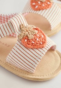 Sandalit valmistatud triibulisest kangast, mille värvitoon on persikane. Rihma peal on heegeldatud oranž maasika dekoratsioon ning sandali all on pruun tekstuuriga tald.