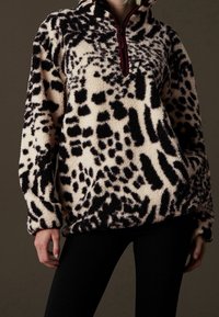 Osoba ubrana w czarny i biały polar z abstrakcyjnym wzorem zwierzęcym oraz bordowym kołnierzem na zamek do połowy długości, zestawiony z czarnymi spodniami.