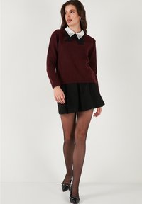 Sieviete, ģērbusies bordo džemperī ar baltu rotātu apkakli un melnu loku, melnās mini svārku, caurspīdīgām zeķbiksēm un melnām norādītām papēžkurpēm, ejot.