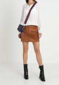 Blusa bianca a maniche lunghe, gonna di velluto a coste marrone con zip frontale, stivali neri lucidi con tacco alla caviglia, borsa a tracolla scura con tracolla a righe.