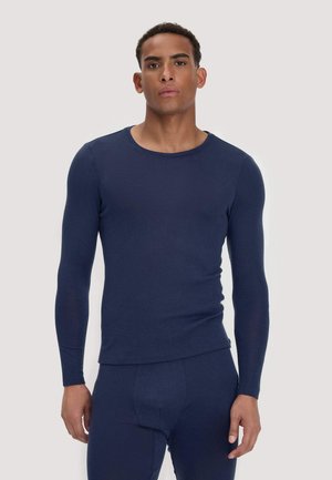 Mann trägt ein eng anliegendes marineblaues Langarmhemd und passende Hose, steht vor einem schlichten hellen Hintergrund.