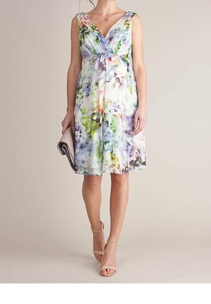 Woman wearing a sleeveless, knee-length floral dress and nude heels, holding a light gray clutch in her left hand.