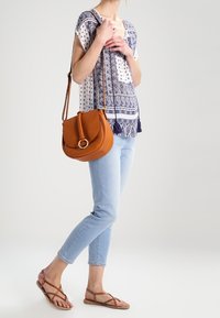 Žena v modrém vzorovaném topu a světlých džínách, s hnědou kabelkou přes rameno a hnědými sandály na pásky, stojící na jedné noze s nohou zvednutou.