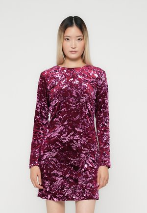 Jonge vrouw met steil ombre haar, gekleed in een korte jurk met lange mouwen bedekt met roze en paarse pailletten, staand tegen een effen achtergrond.