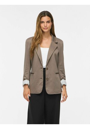 Kvinde med langt brunt hår iført en taupefarvet blazer over en hvid top og sorte bukser står mod en ensfarvet hvid baggrund.