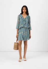 Robe à imprimé floral en tissu bleu-vert doux, avec un col froncé et des manches trois-quarts, accompagnée d'un sac rond en toile et de sandales.