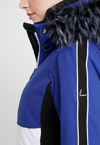 Gros plan d'une personne portant une veste bleu et blanc avec une capuche bordée de fourrure noire et une bande verticale noire avec un emblème métallique en forme de "L".