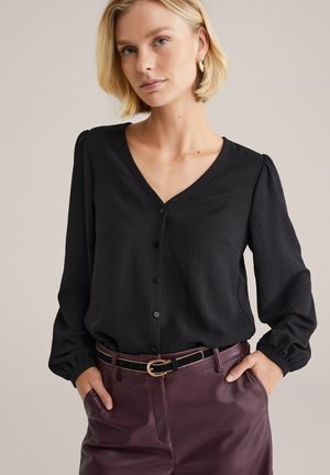 Blondynka w czarnej zapinanej na guziki bluzce z długimi rękawami, wpuszczonej w wysokowalne burgundowe skórzane spodnie z czarnym paskiem.