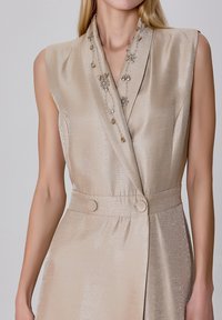 Robe sans manches beige avec un décolleté en V, ornée de perles et de sequins, présentant une taille cintrée et des accents de boutons.