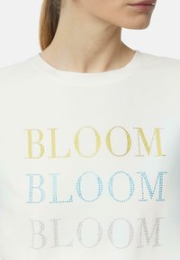 Witte t-shirt met korte mouwen, met de tekst "BLOOM" in gele, blauwe en zilveren rhinestones, voorzien van een klassieke ronde hals en een zachte stoftextuur.
