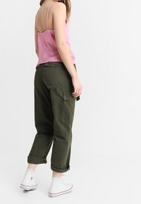 Donna che indossa un top rosa a spaghetto, pantaloni cargo verde scuro con risvolti e sneakers bianche alte, in piedi di lato.